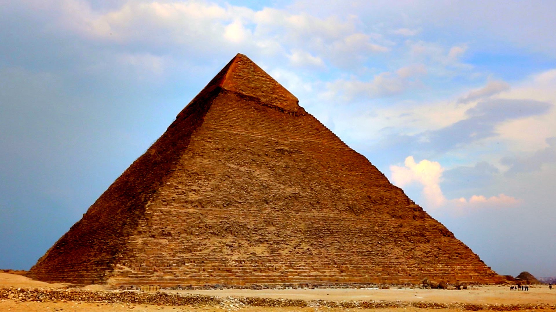 pirâmide com algumas pessoas no canto direito inferior, e no fundo o céu azul com algumas nuvens e manchas rosas